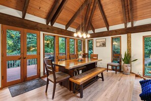 Comedor con mesa de madera, sillas y bancos, ventanas amplias con vista a follaje y estructura de techo de madera.