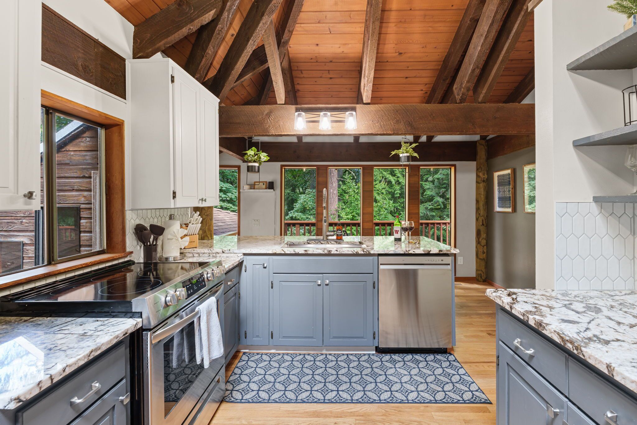 A modern kitchen with a central island, stainless steel appliances, and a view of the forest through large windows.