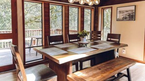 A wooden dining table with six chairs, a bench, and a potted plant. Large windows allow natural light and views of trees.