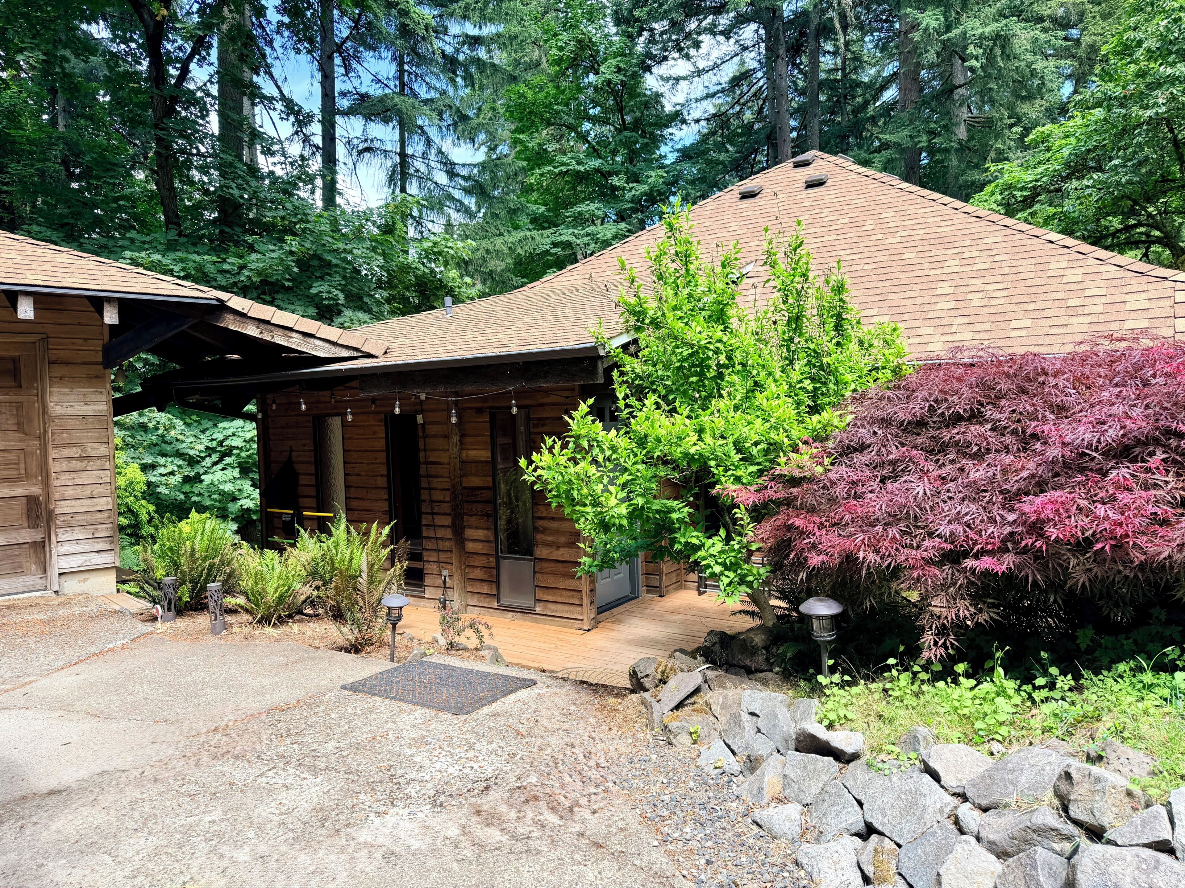 A wooden cabin with a covered porch, surrounded by dense forest and a gravel driveway.