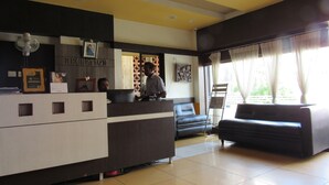 A reception area with a reception desk, a sofa, and a window with curtains. - Devis Grand (Puducherry)