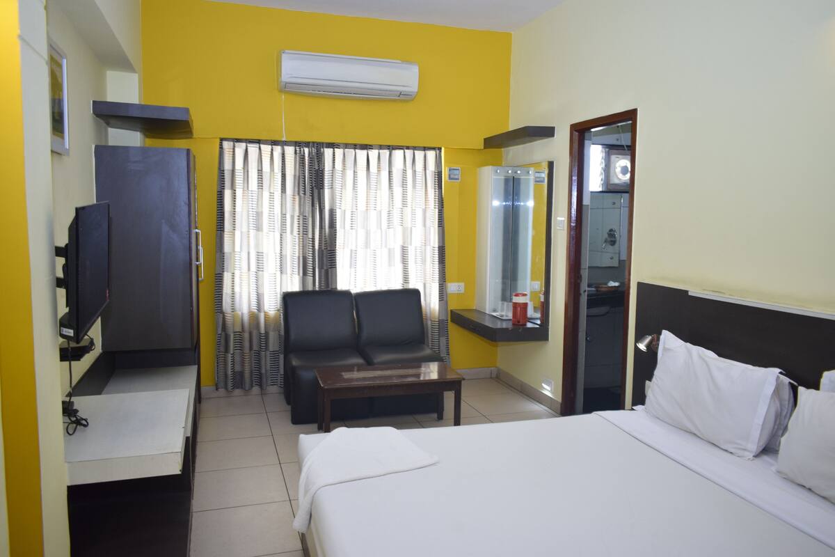 A hotel room with a bed, a desk with a chair, a television, a sofa, and a window with curtains.