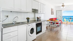 A modern kitchen with white cabinets, a built-in washing machine, and a dining area with a red sofa and a view of the sea. - 1.1 Beautiful seafront with balcony and WIFI (Arona)