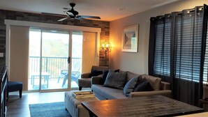 Een woonkamer met een bank, salontafel en een open haard. Er is een schuifdeur die leidt naar een balkon met tuinmeubilair.