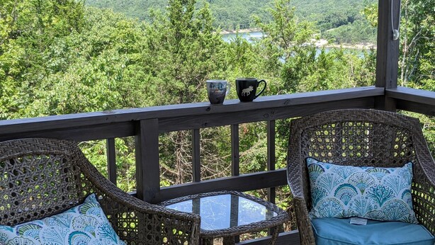 A wicker chair with a blue cushion and a glass table on a wooden deck with a railing, overlooking a forested valley.