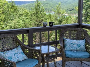 A wicker chair with a blue cushion and a glass table on a wooden deck with a railing, overlooking a forested valley. - Canaan Point (Branson West)