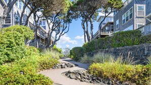 A pathway leading through a landscaped area with dense greenery and trees, flanked by multi-story buildings with balconies. - Wonderful Coastal Retreat, Open Layout, Steps Away From Beach, Fireplace, BBQ, Ocean Views (Depoe Bay)