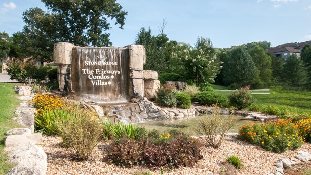 A stone sign with a waterfall feature, surrounded by a landscaped garden with various plants and flowers. - The Plush (Branson West)
