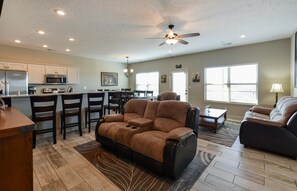 A spacious living area with a large sofa, a dining table with chairs, a kitchen with modern appliances, and a ceiling fan.