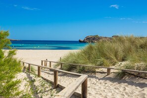 A sandy beach with a wooden walkway leading to the water, surrounded by dunes and sparse vegetation. - Apartment Pregonda 13 Menorca with Wi-Fi, Pool, Terraces & Garden (Son Parc)