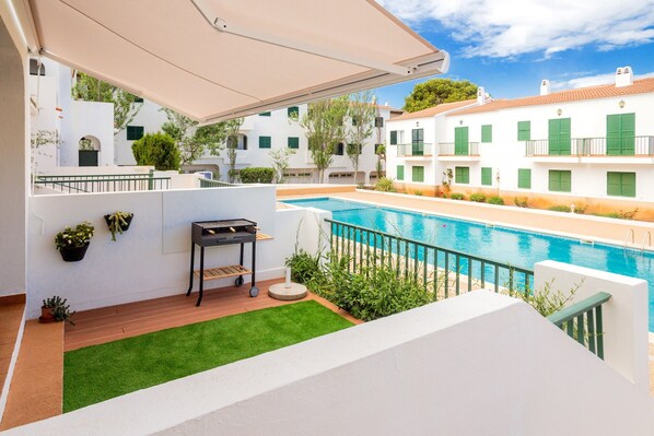 A poolside area with a grill, potted plants, and a view of white buildings with green shutters. - Apartment Pregonda 13 Menorca with Wi-Fi, Pool, Terraces & Garden (Son Parc)