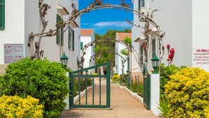 Una entrada cerrada con un arco verde, edificios blancos con contraventanas verdes y abundante vegetación.