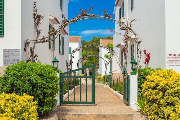 A gated entrance with a green archway, white buildings with green shutters, and lush greenery.