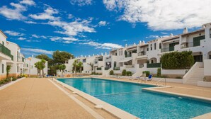 Zona de piscina con cielo despejado, edificios blancos y un paseo marítimo de arena.