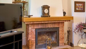A brick fireplace with a clock and decorative items on the mantel, a flat-screen TV on a stand, and a glass vase with white pebbles on a table.