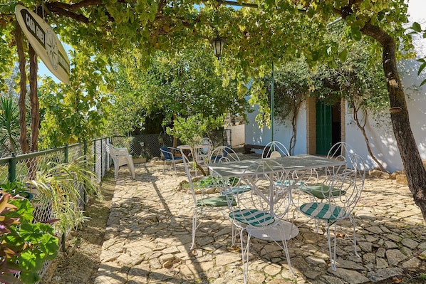 An outdoor patio with stone flooring, metal chairs, and tables, surrounded by greenery and a white building with a green door.