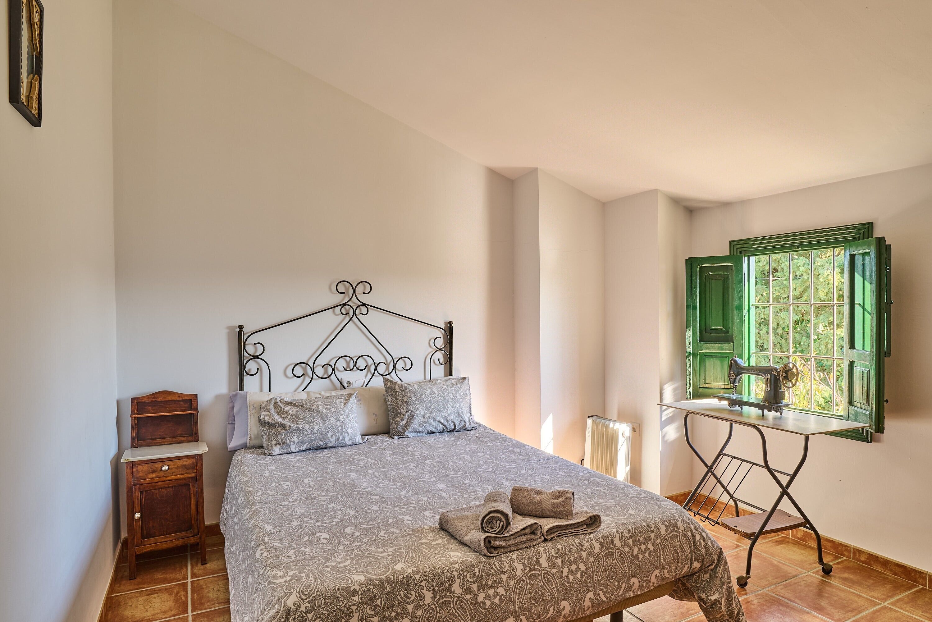 Una camera da letto con un letto, un comodino, un tavolo da cucito e una finestra con persiane verdi.