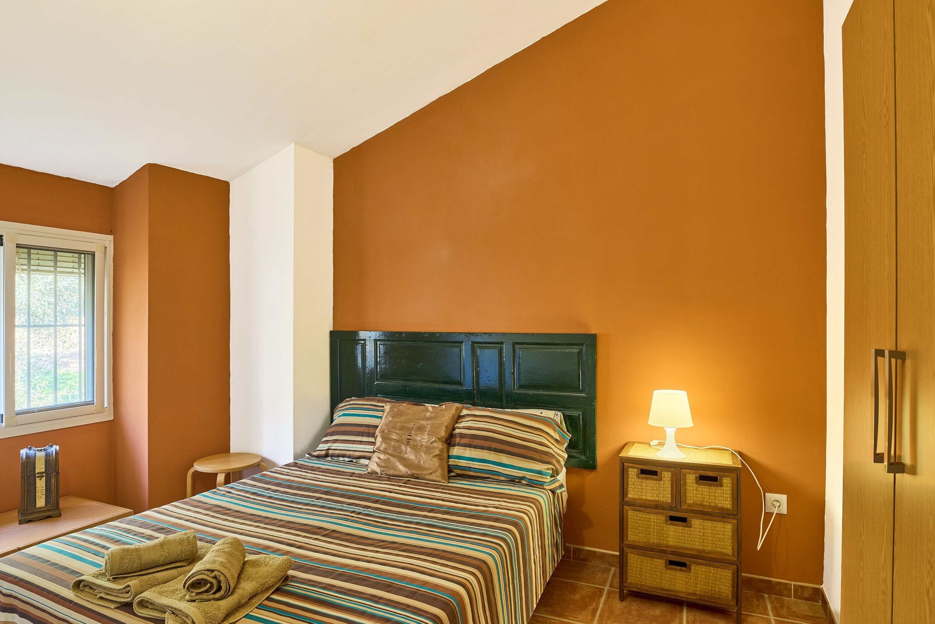 A bedroom with a bed, a bedside table with a lamp, a small stool, and a cabinet.