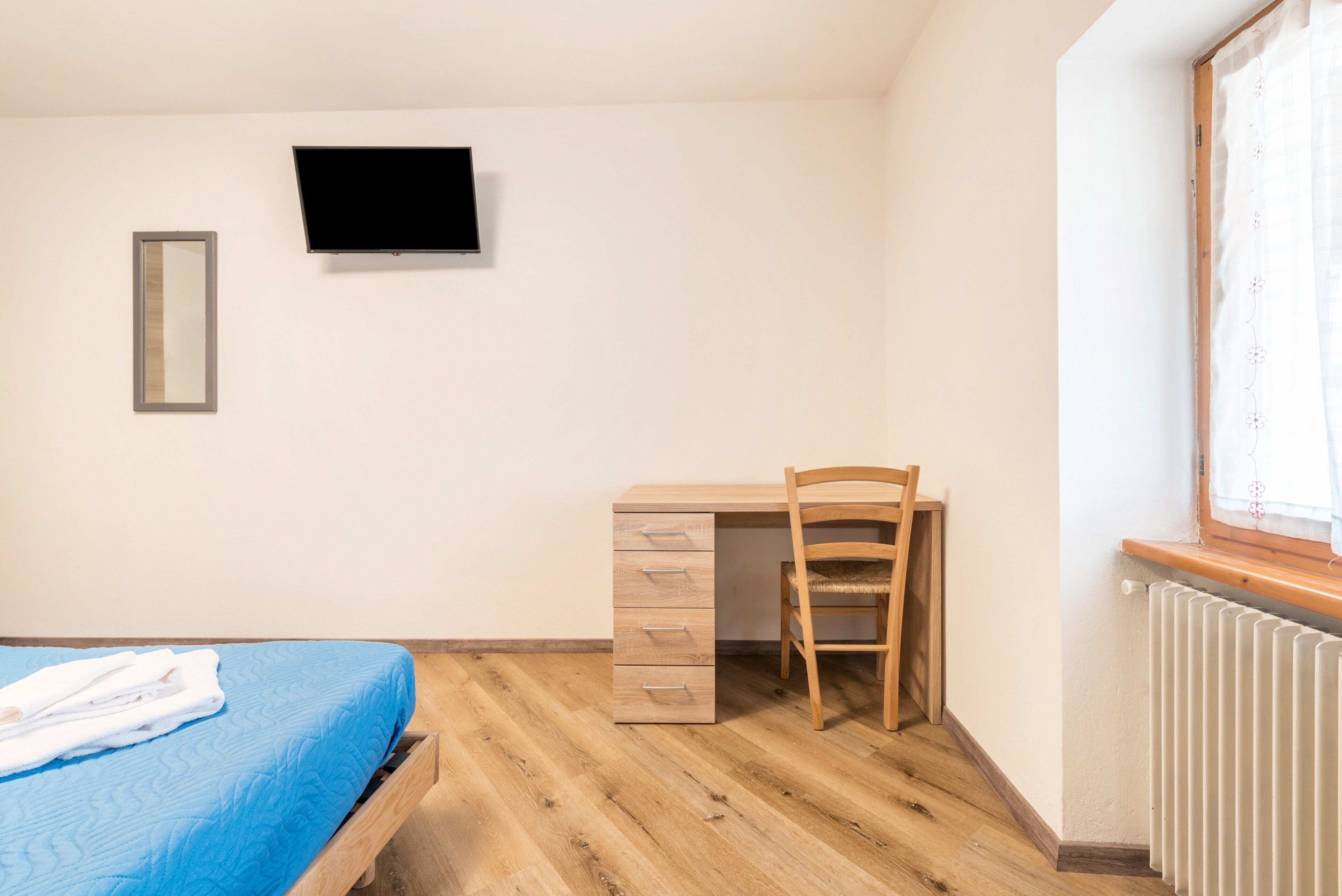 A bedroom with a bed, a desk, a chair, a TV, and a window.