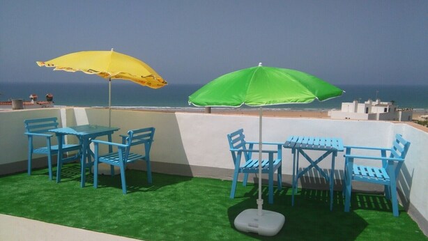 A rooftop terrace with blue chairs and tables, two umbrellas, and a view of the sea.