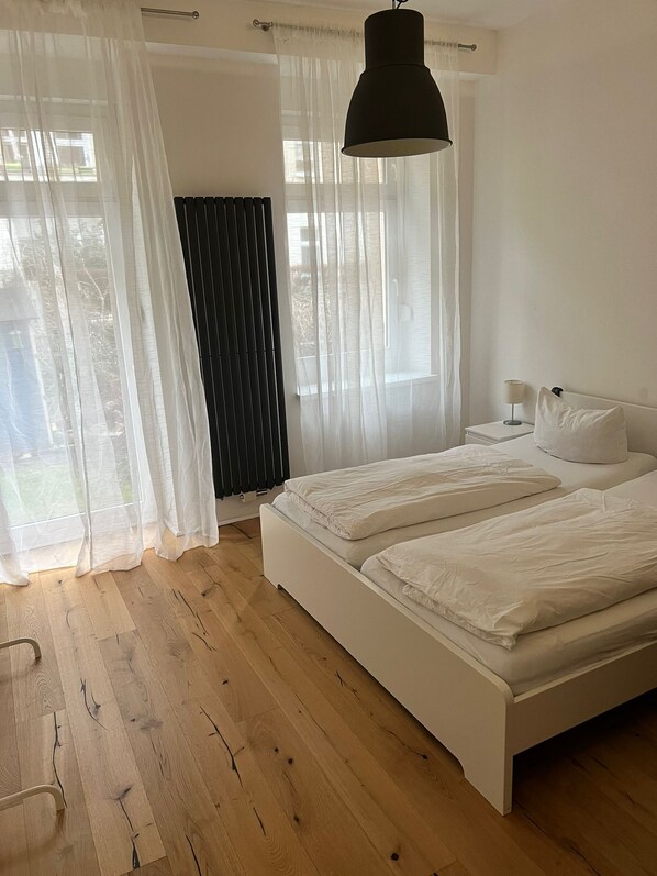 A bedroom with a large bed, a bedside table with a lamp, sheer curtains, and a view of the outdoors.