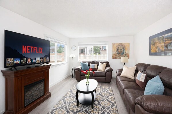 A living room with a fireplace, a television displaying Netflix, a brown leather sofa, a coffee table, and a patterned rug.