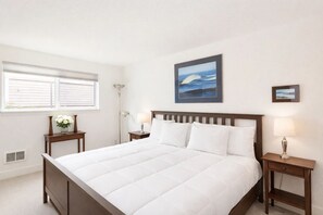 A neatly made bed with white bedding, a wooden headboard, and two bedside tables with lamps. There is a window with blinds, a wall-mounted picture of the ocean, and a vent on the wall.