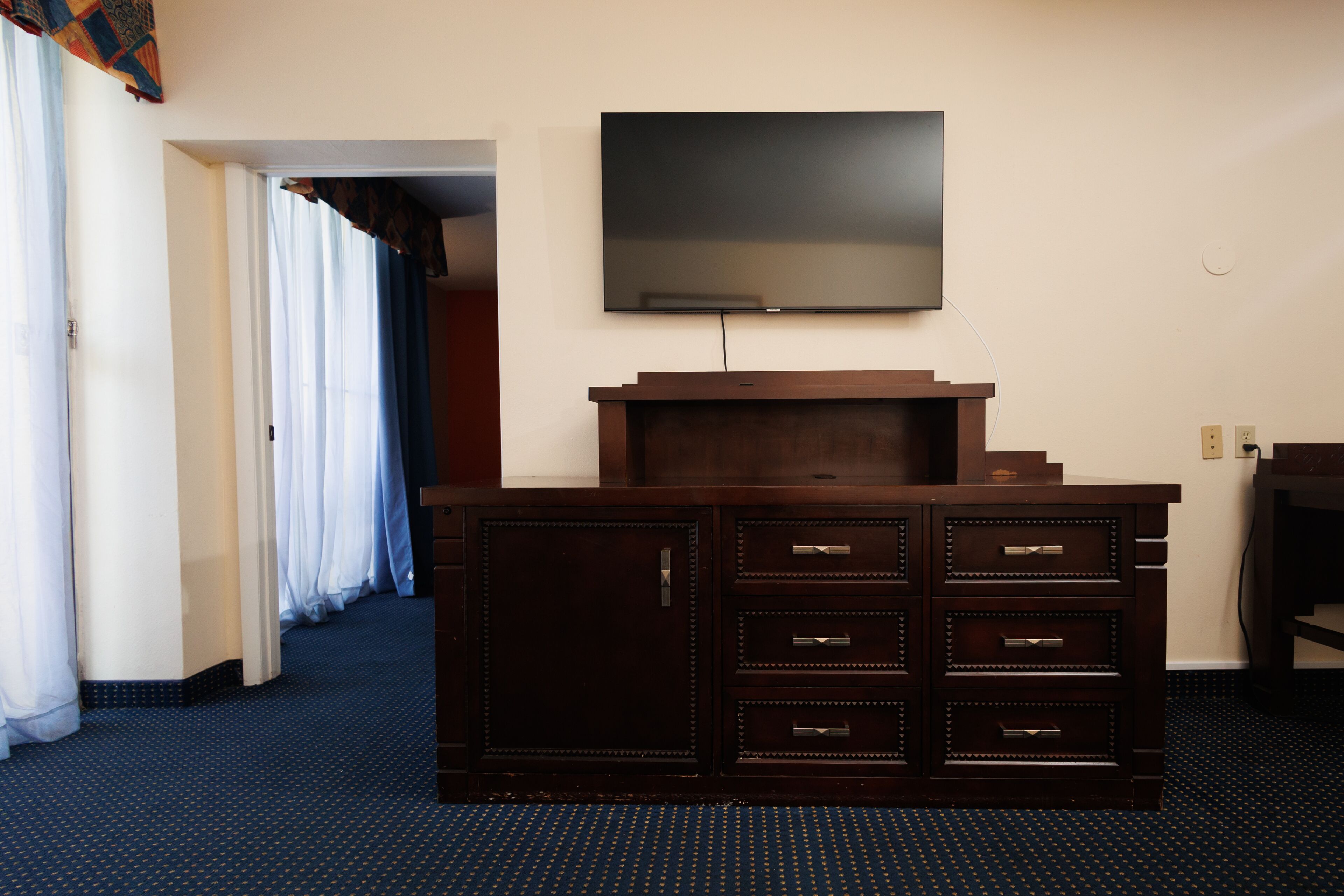 A hotel room with a dark wooden dresser, a flat-screen TV mounted on the wall, and a blue carpeted floor.