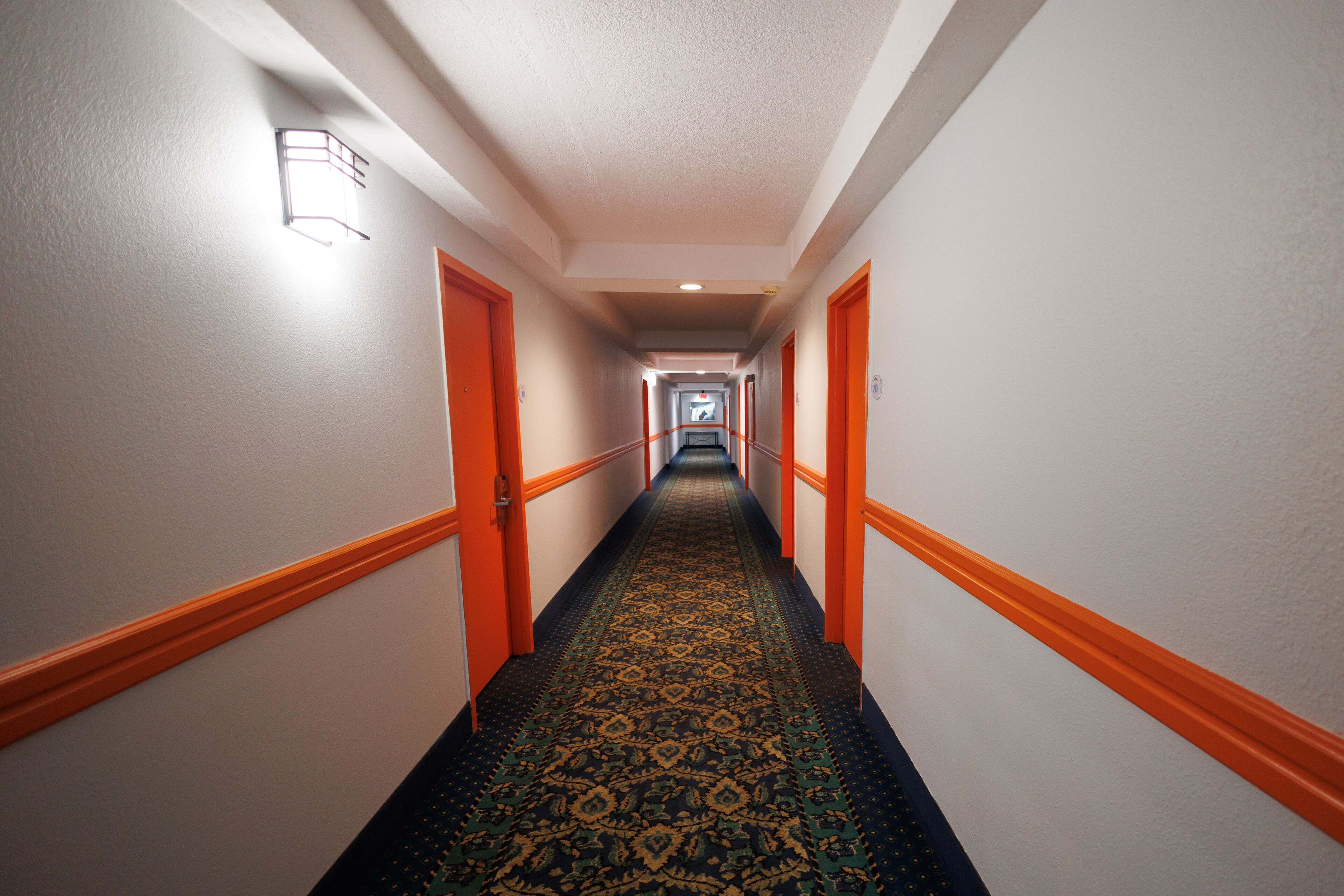 A hotel hallway with a patterned carpet, orange doors, and a light fixture on the wall.