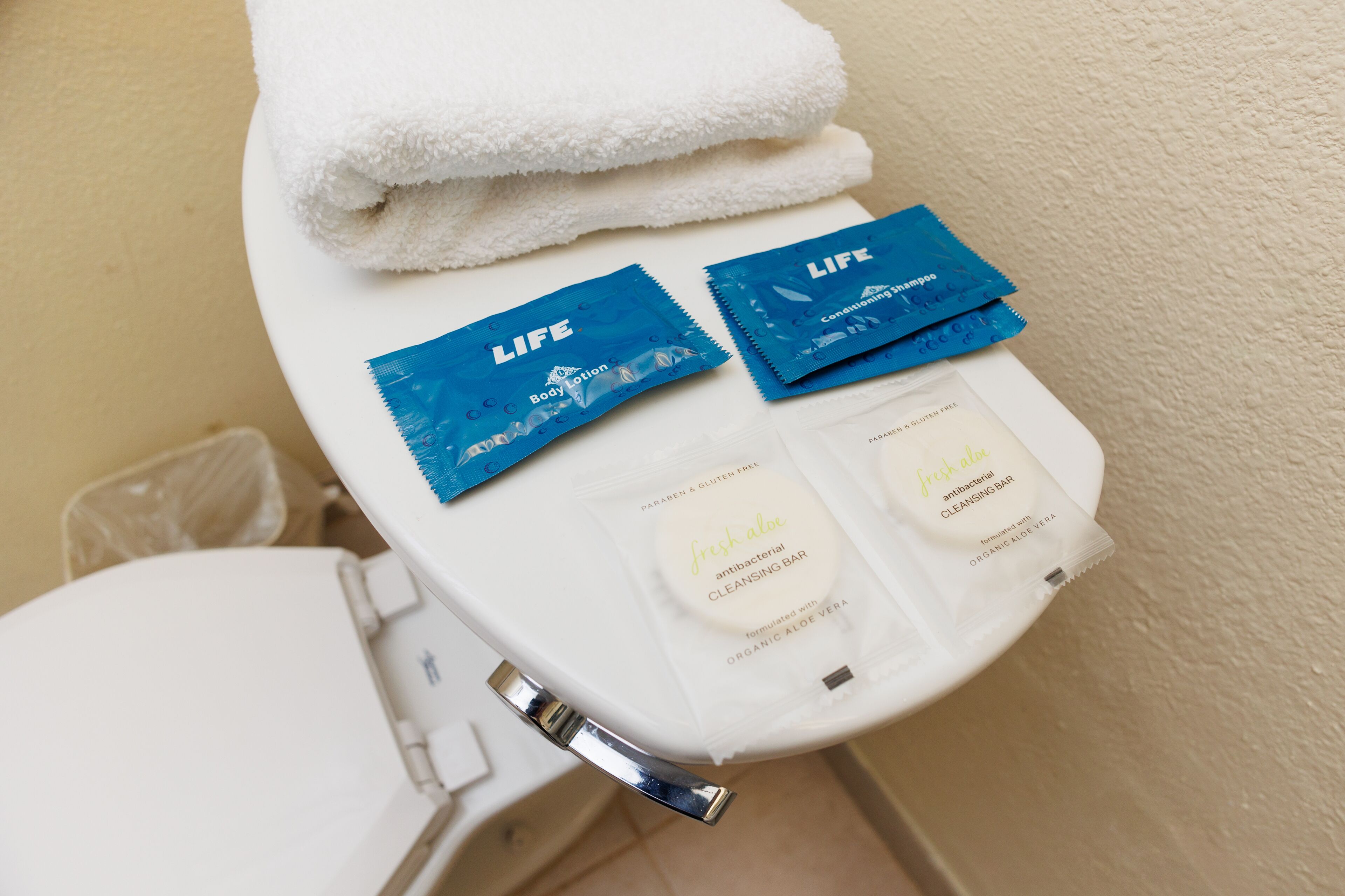 A bathroom with a white toilet, a stack of white towels, and two blue Life Body Lotion packets and a fresh aloe antibacterial cleansing bar on the toilet seat.