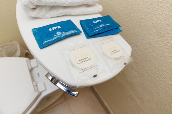 A bathroom with a white toilet, a white towel on the seat, and two blue Life Beauty Lotion packets and a Fresh Aloe Cleansing Bar packet on the lid. - Garnet Inn & Suites, Orlando (Orlando)