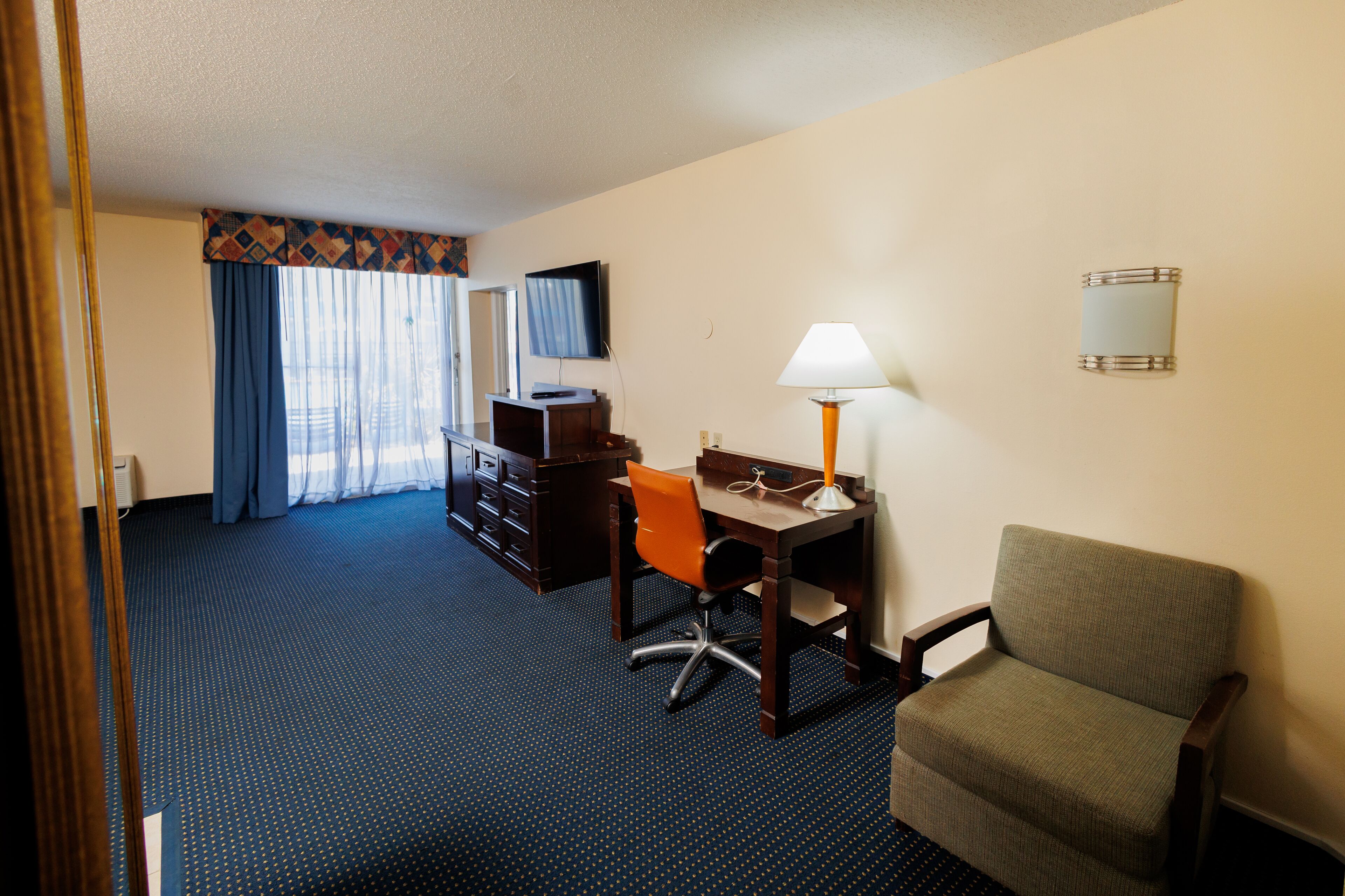 A hotel room with a desk, chair, lamp, TV, and a bed with a blue curtain.