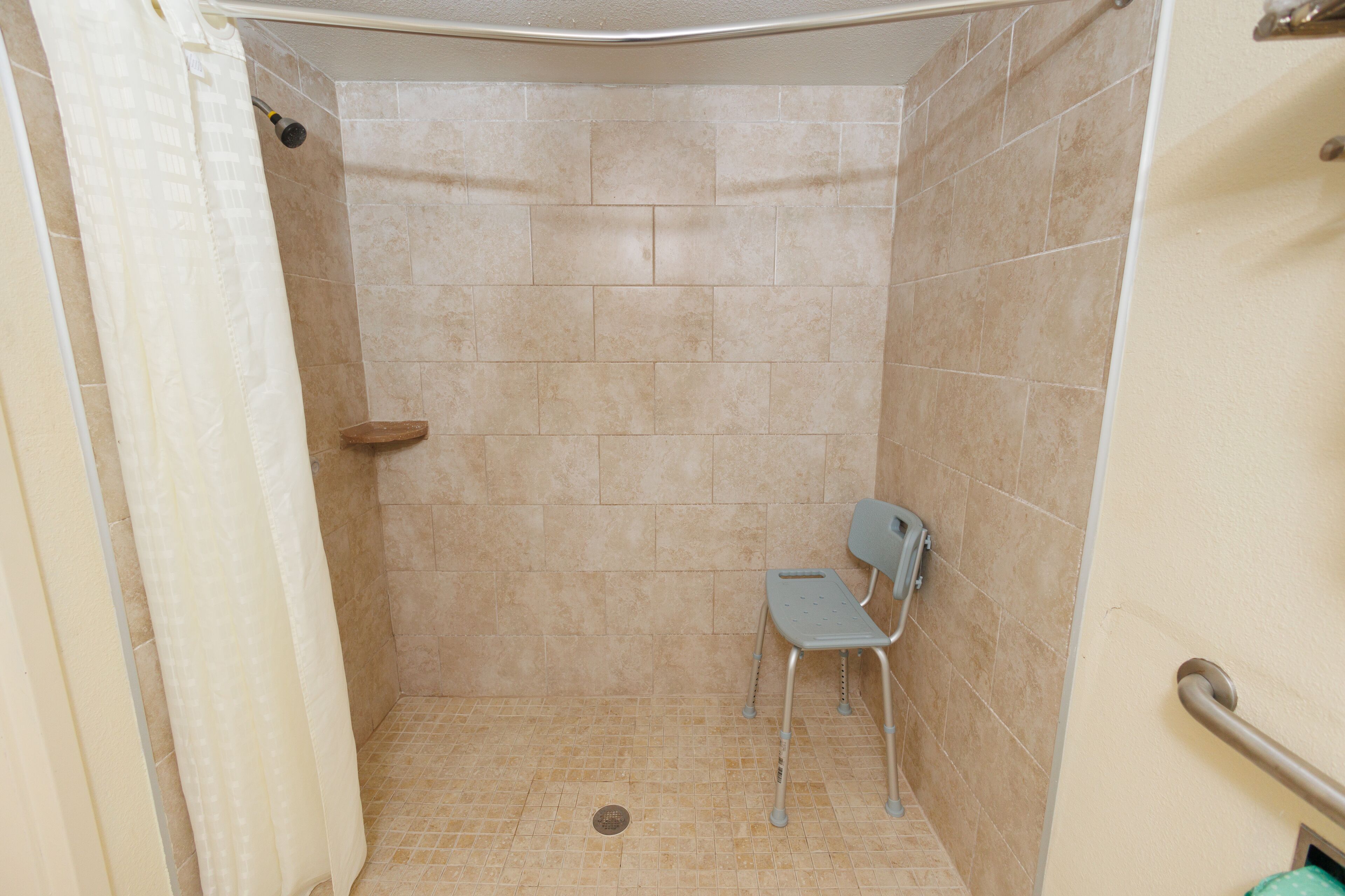 Un box de ducha con azulejos color beige, un cabezal de ducha, un asidero y un asiento accesible para sillas de ruedas.
