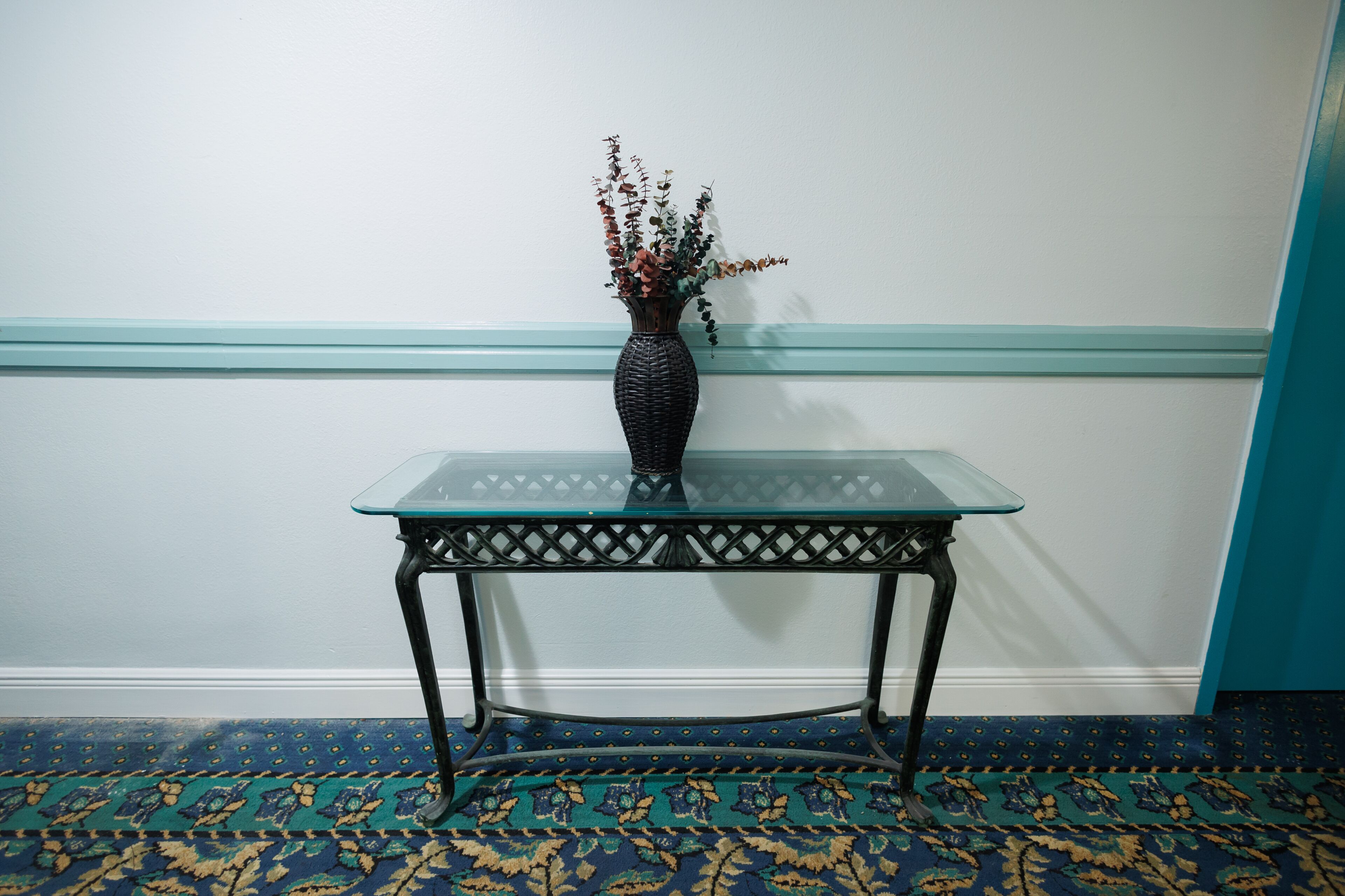 Una mesa con superficie de vidrio, un jarrón tejido con flores secas, ubicada frente a una pared con detalles azul turquesa y una alfombra estampada.