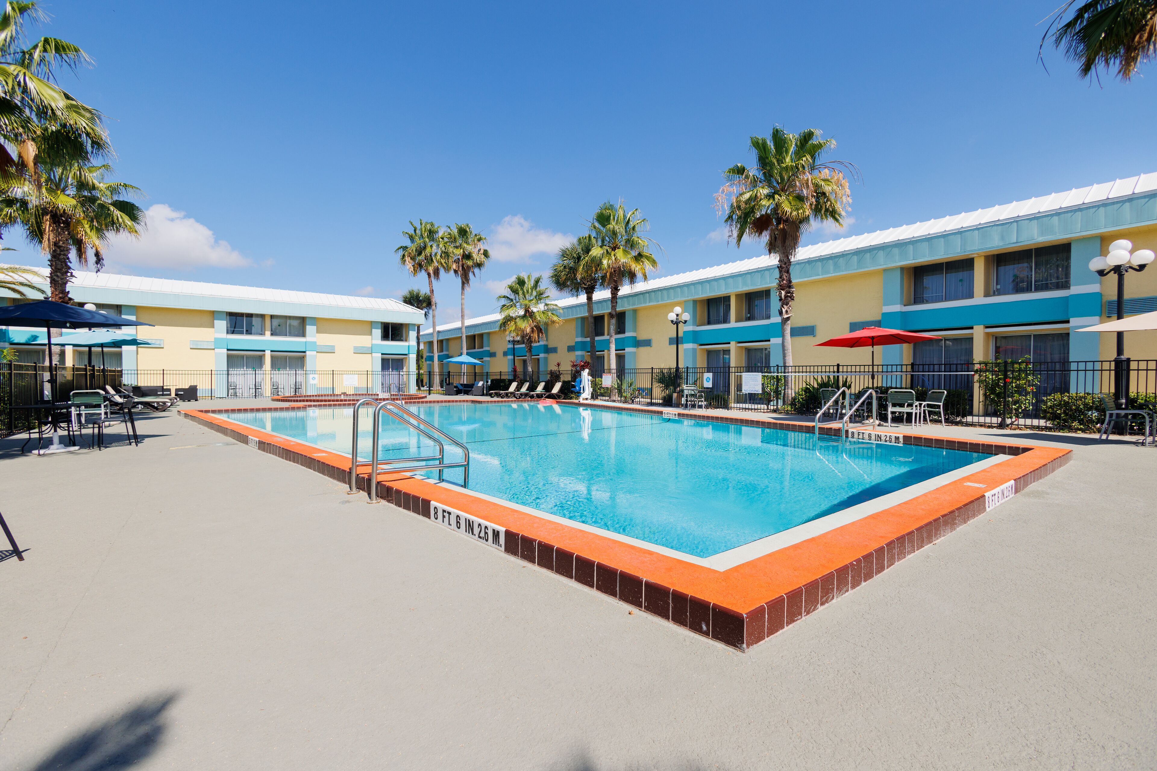 A swimming pool with a blue and orange border, surrounded by palm trees and a hotel building with blue and yellow exterior walls.