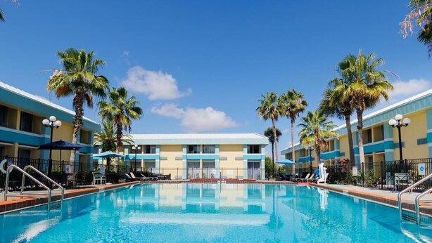 A swimming pool with lounge chairs and umbrellas, surrounded by palm trees and a hotel building with a blue and yellow facade. - Garnet Inn & Suites, Orlando (Orlando)