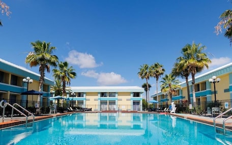 A swimming pool with lounge chairs and umbrellas, surrounded by palm trees and a hotel building with a blue and yellow facade.