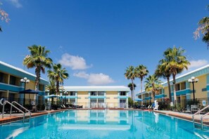Uma piscina com espreguiçadeiras e guarda-sóis, cercada por palmeiras e um edifício de hotel com fachada azul e amarela.