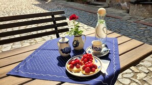 Una mesa al aire libre con mantel azul, un plato de fresas, una jarrón y una tetera, y una rosa.