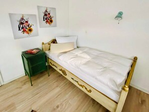 Una cama bien tendida con estructura de madera y ropa blanca, una mesita de noche pequeña de color verde con un libro, y dos cuadros decorativos con motivos florales.