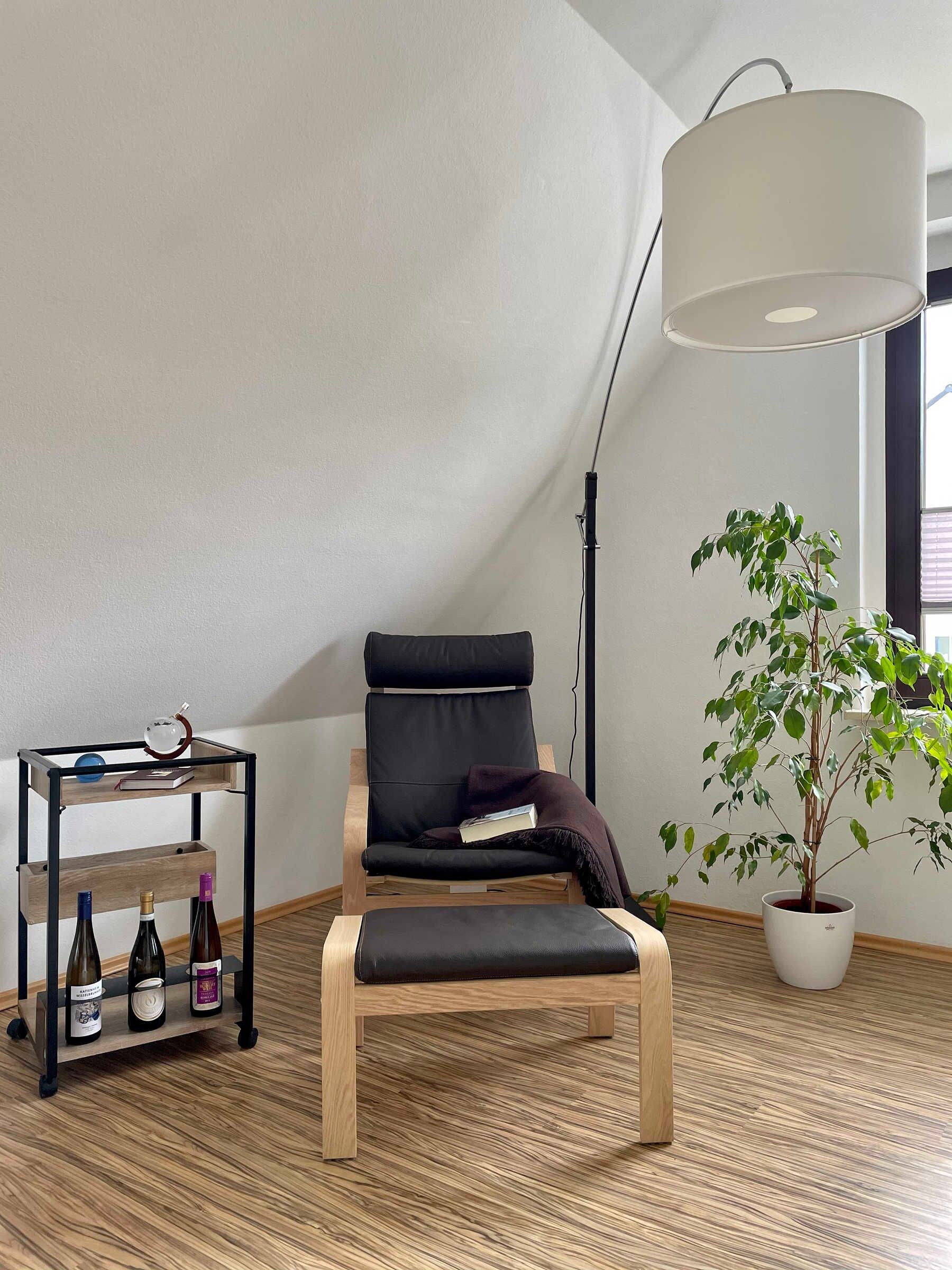 A modern room with a black leather chair, a wooden floor, a small table with bottles, a potted plant, and a large lamp.