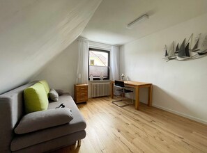 A small, well-lit room with a wooden floor, a grey sofa with a green pillow, a wooden desk, and a radiator. - Hänsel Vacation Rental, Germany (Zinnowitz)
