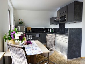 A modern kitchen with dark cabinetry, a dining area with a wicker chair, and a table set with flowers and glasses. - Hänsel Vacation Rental, Germany (Zinnowitz)