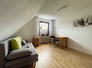 A small, well-lit room with a wooden floor, a grey sofa with green cushions, a wooden desk, and a radiator. - FeWo Hänsel, Germany (Zinnowitz)