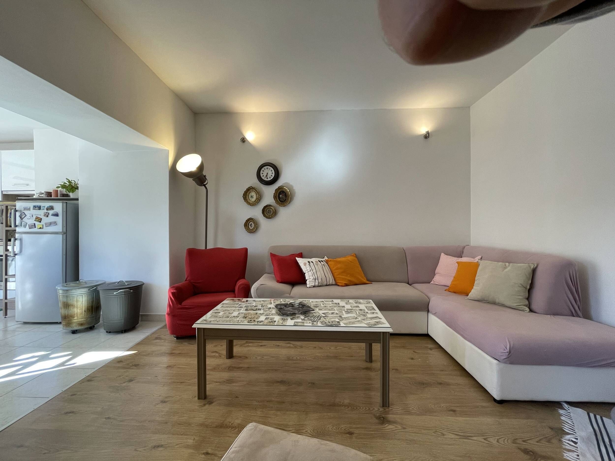 A modern living room with a sofa, a red armchair, a coffee table with newspapers, and a wall clock.