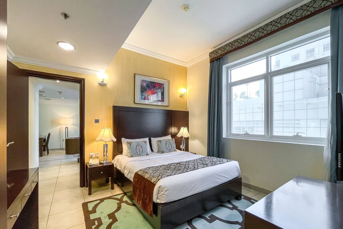 A hotel room with a bed, bedside lamps, a nightstand, a desk, a window, and a door leading to another room.