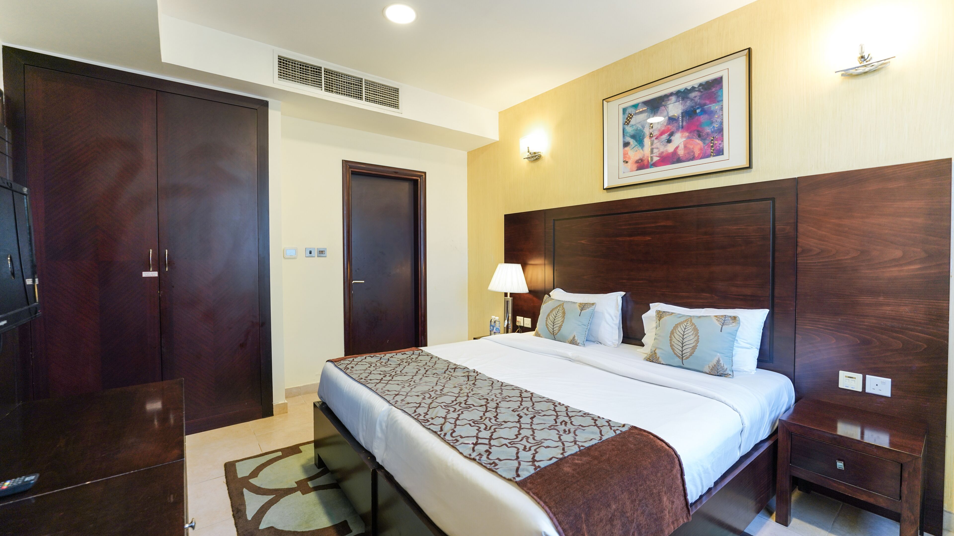 Quarto de hotel com uma cama grande, um guarda-roupa, uma mesa de cabeceira com abajur e uma obra de arte emoldurada na parede.