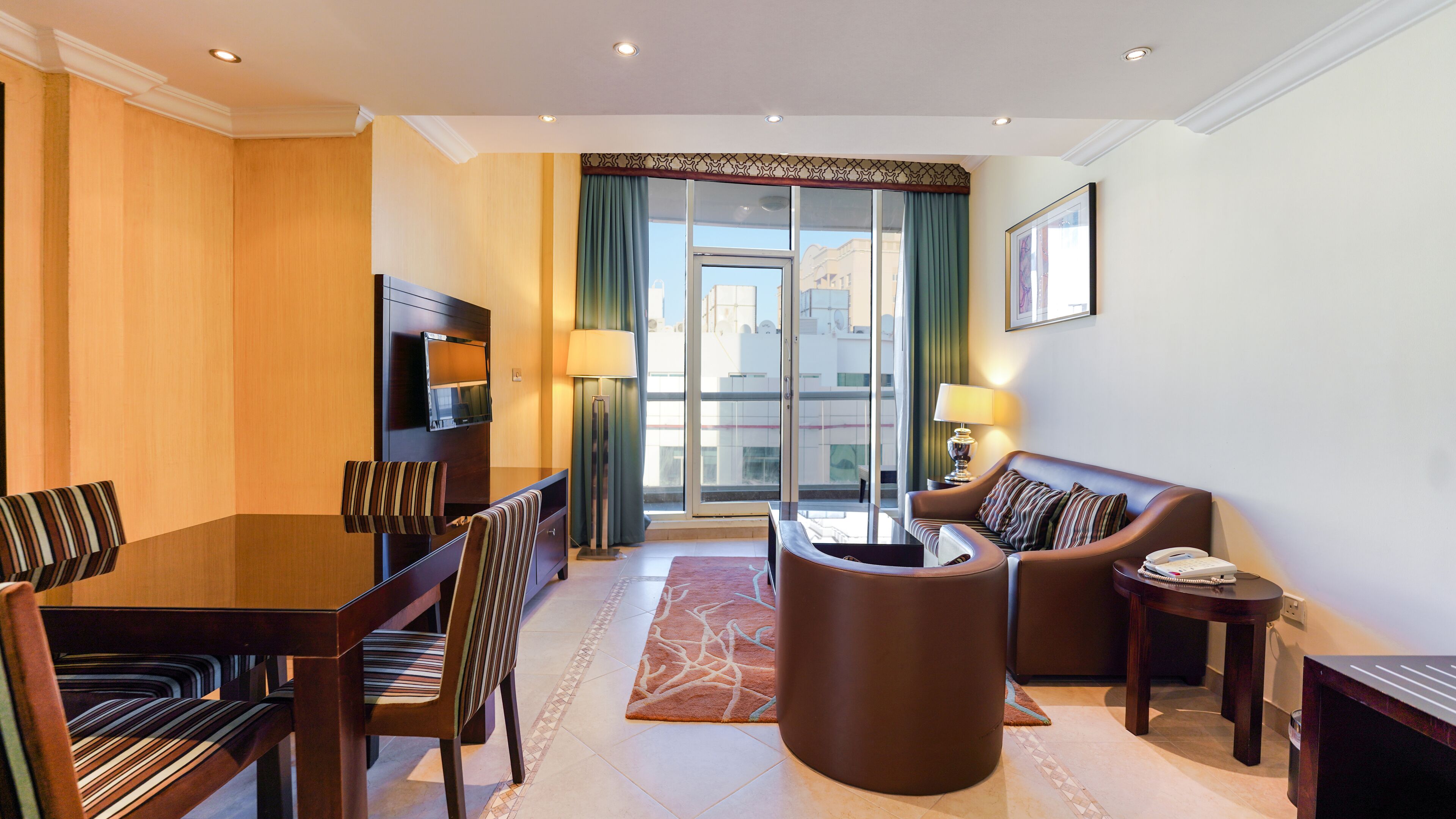 Una habitación de hotel moderna con una mesa de comedor, dos sofás, un televisor y un ventanal con cortinas.