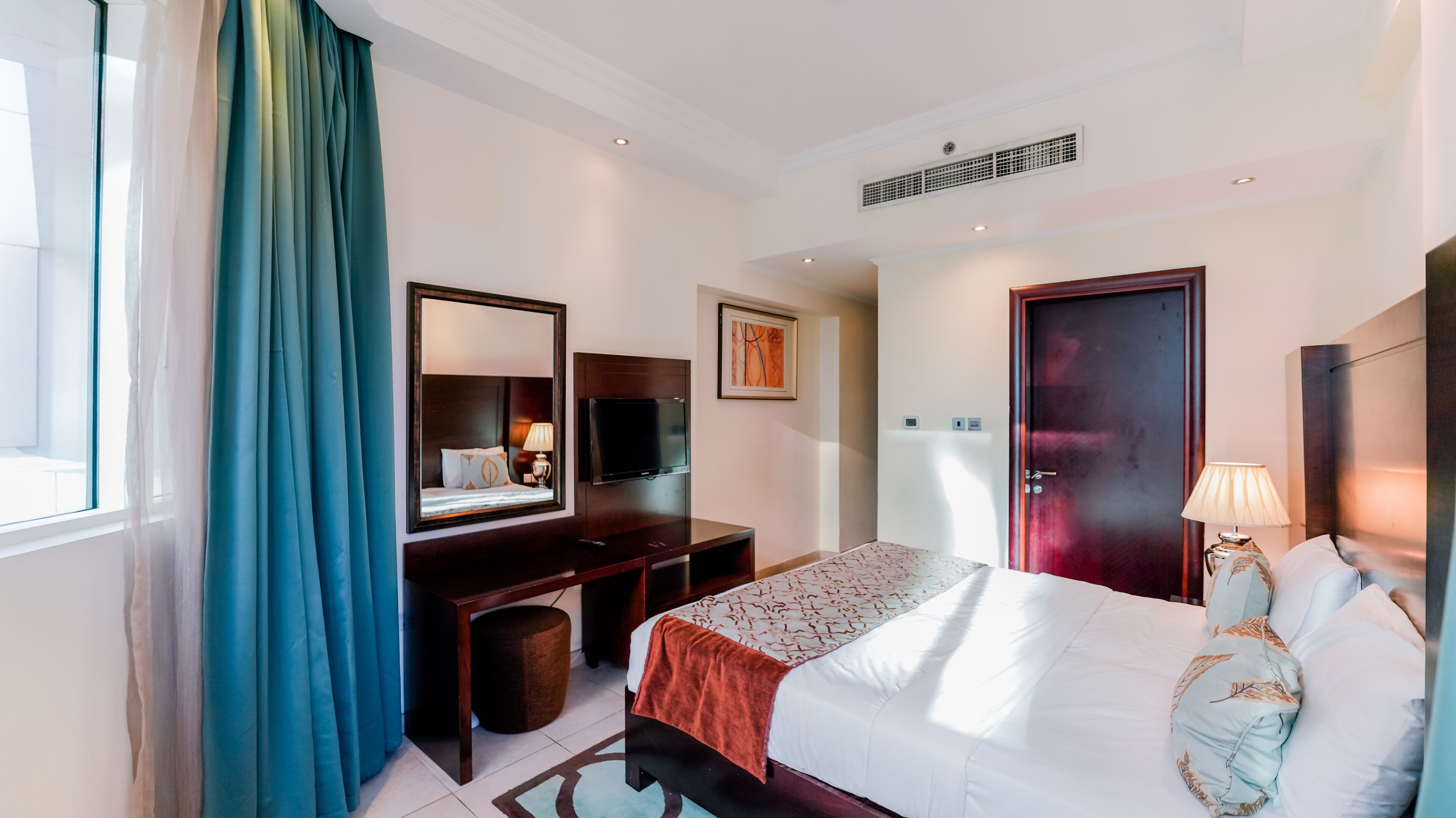 Ein Hotelzimmer mit einem Bett, einem Schreibtisch mit Fernseher, einem Spiegel, einer Tür und einem Fenster mit Vorhängen.