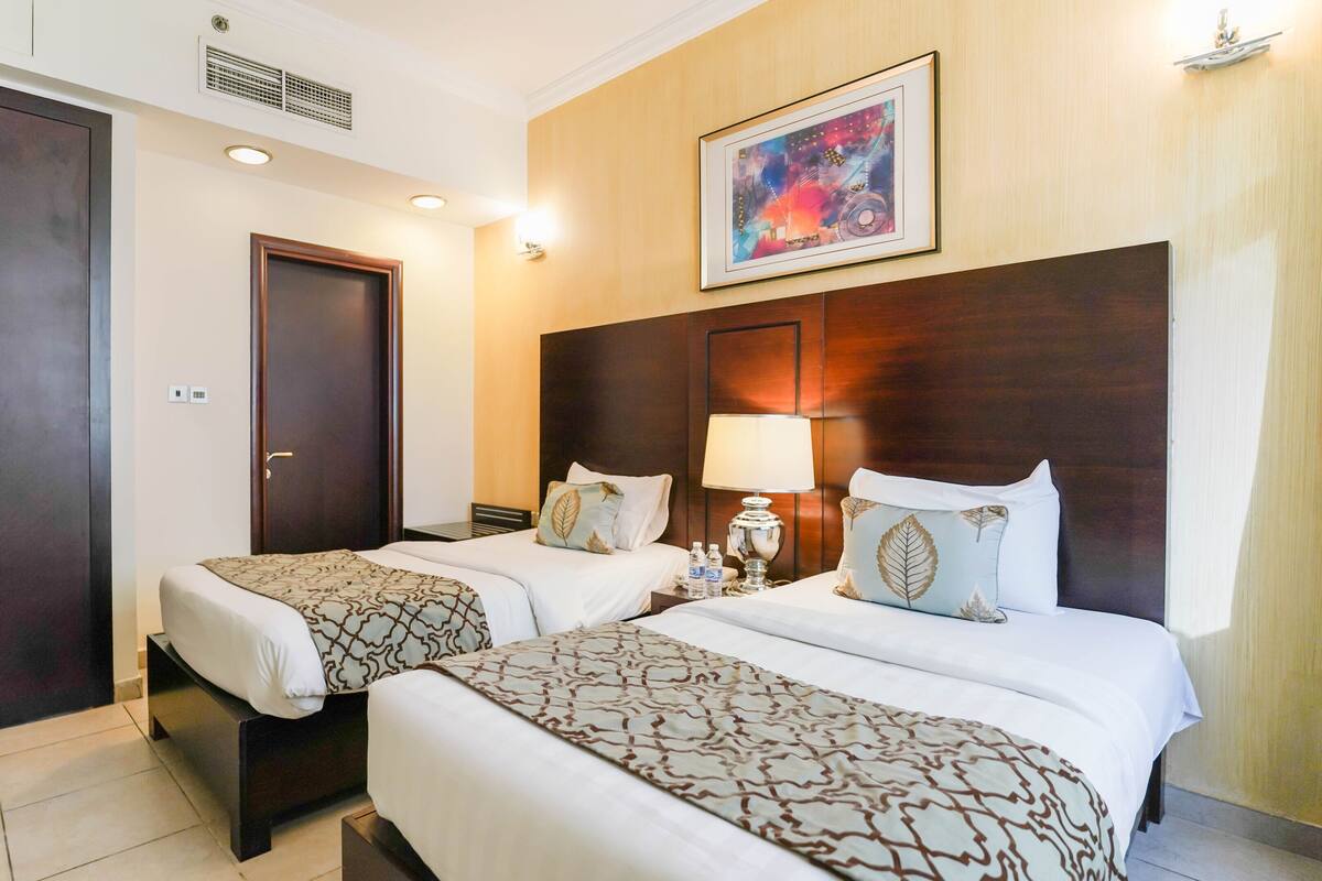 A hotel room with two beds, a large wardrobe, a painting on the wall, and a bedside table with a lamp.